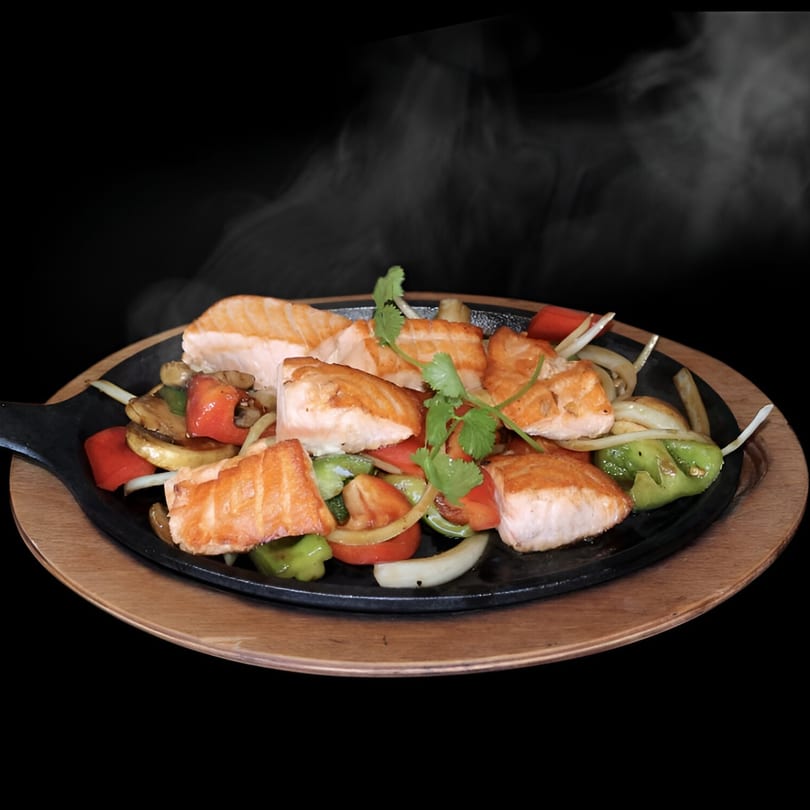 Steaming hot salmon and stir-fried vegetables on a sizzling black plate with a wooden base.