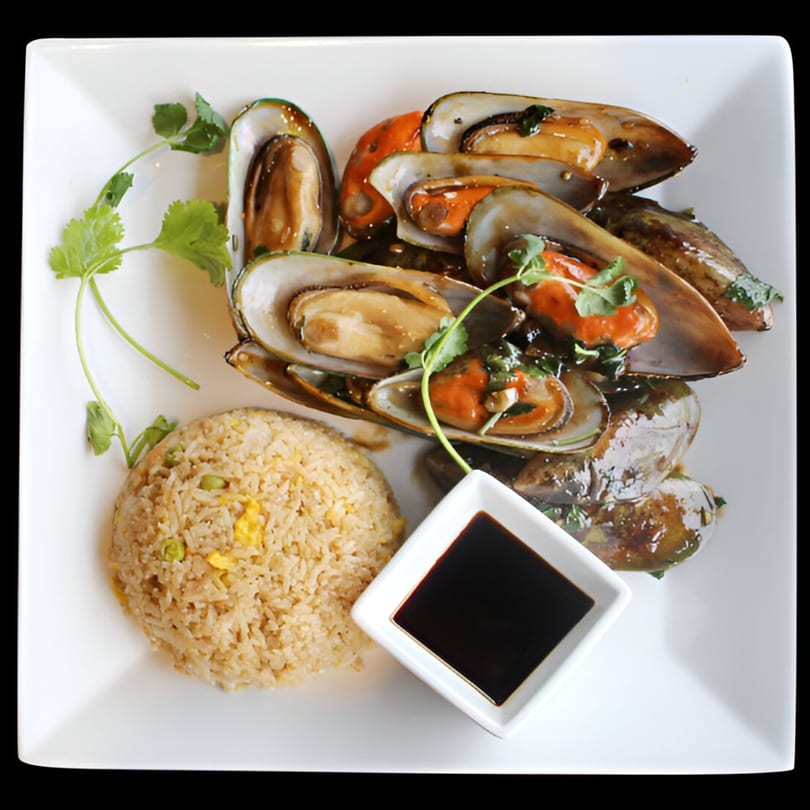 Mussels in a savory sauce with fried rice and a side of soy sauce on a white plate.