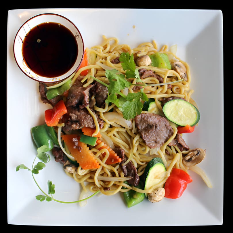 Vietnamese beef stir-fried noodles with colorful vegetables and a side of dark sauce.