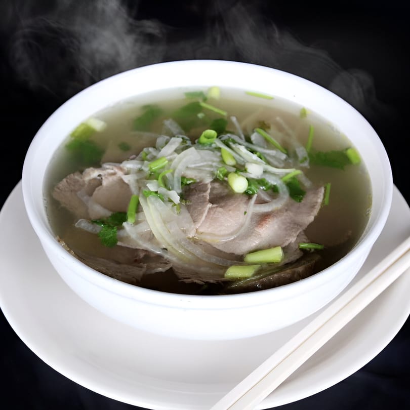 Steaming bowl of Vietnamese pho with beef, onions, and green herbs, served with chopsticks.